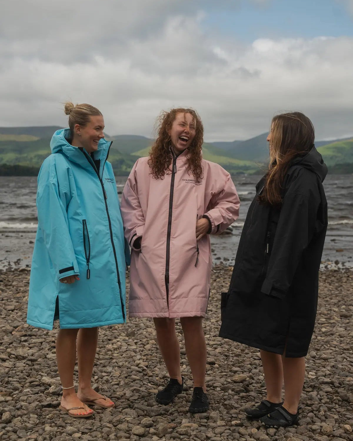 Three women standing on a pebble beach wearing oversized waterproof ColdWaterCO raincoats in light blue, dusty pink, and black, with hills and a cloudy sky in the background.