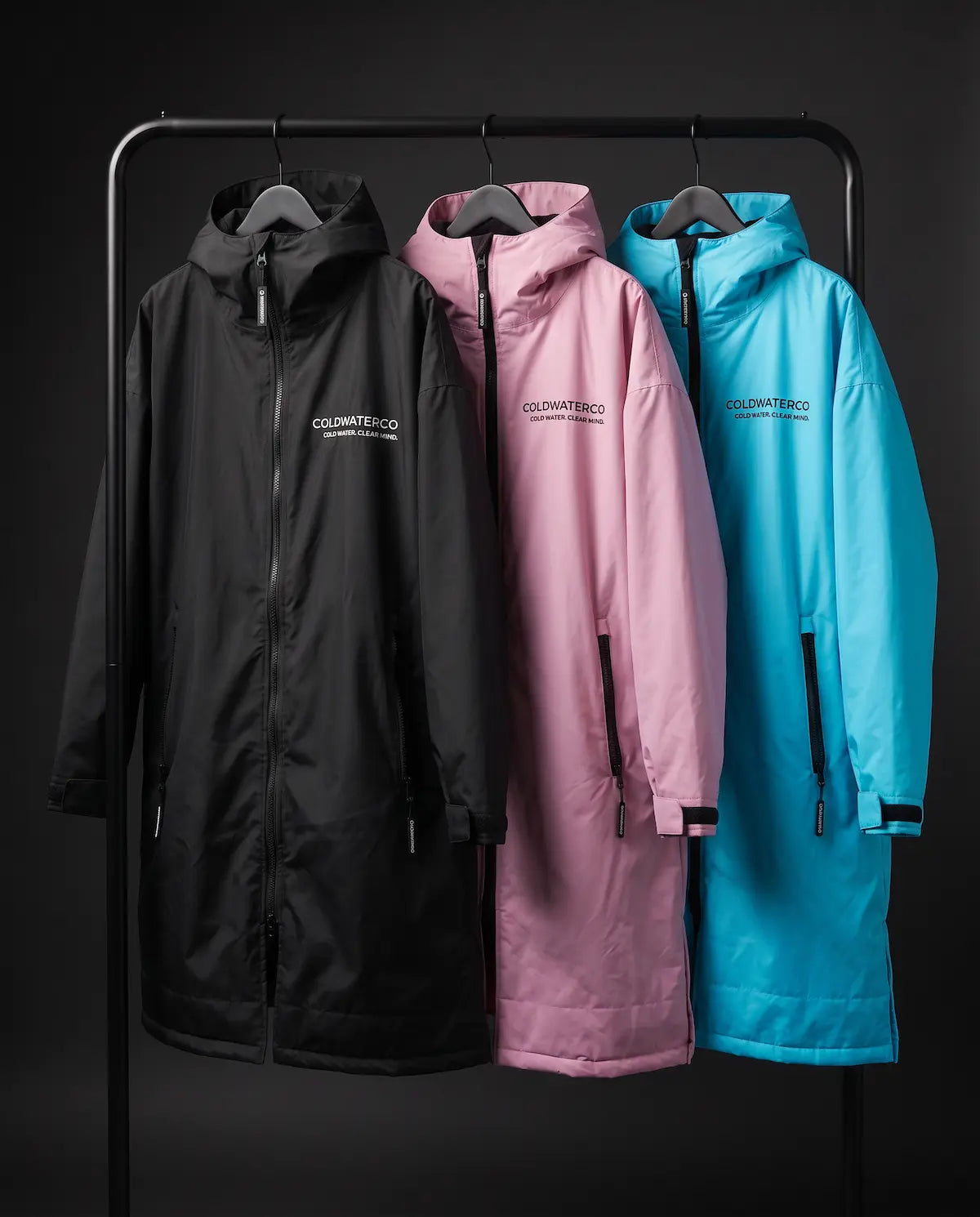 Three hooded waterproof changing robes hang on a black clothing rack against a dark studio background: one black, one dusty pink, and one bright turquoise blue. Each coat has a front zipper, side zippers, and a small white 'COLDWATERCO' logo on the chest.