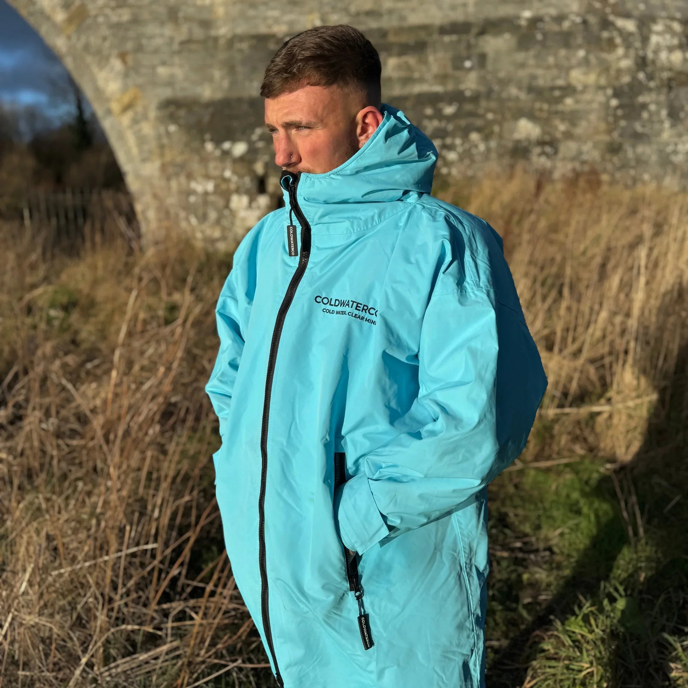 Person wearing a light blue ColdWaterCo jacket with a brand logo, standing outdoors near a stone bridge.