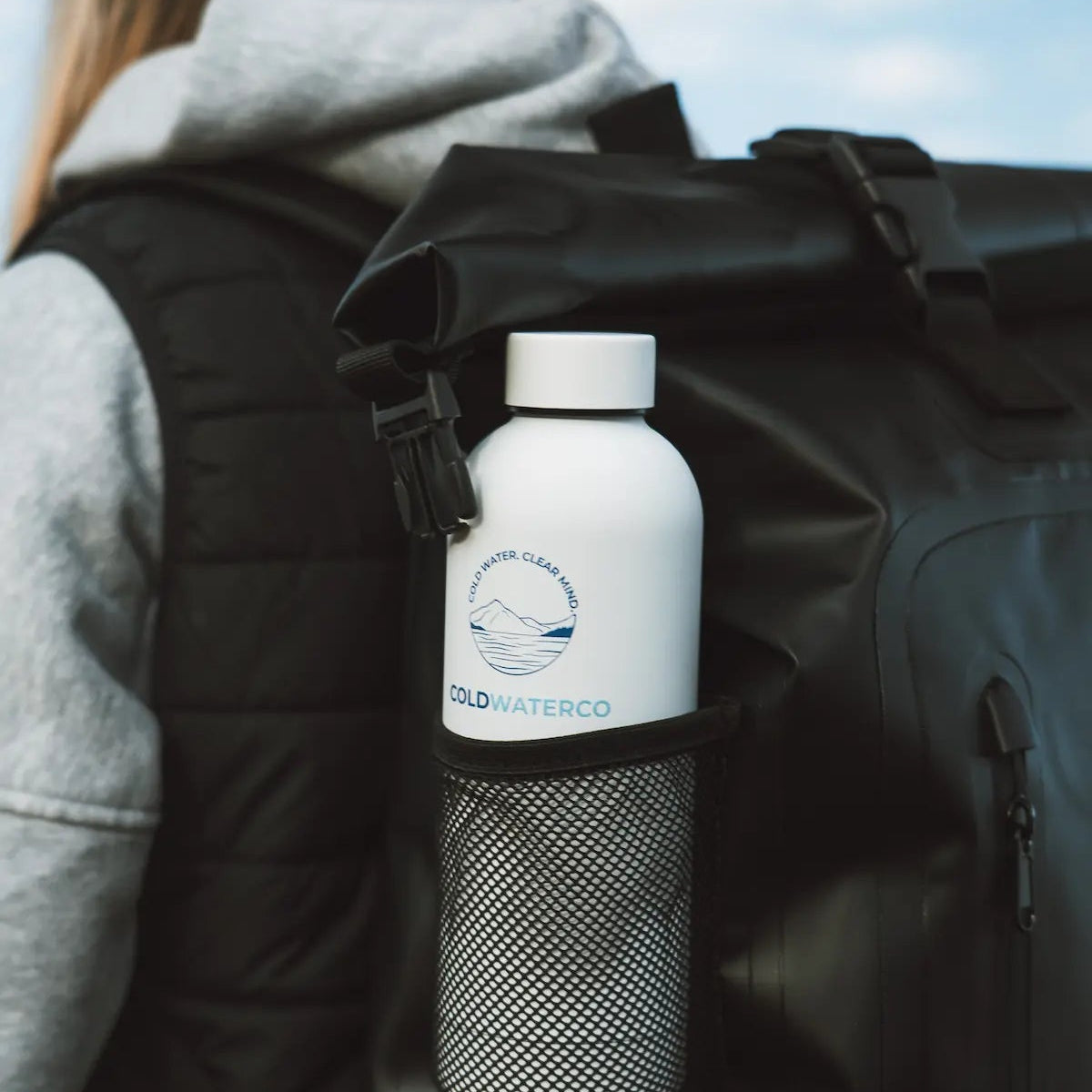 Person with a black backpack holding a white water bottle labeled 'Cold Water Co.' against a blue sky.