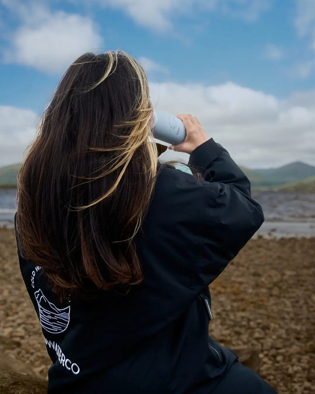 A woman with long brown hair with blonde streaks is seen from behind, wearing a black COLDWATERCO jacket with a white graphic on the back and holding a white COLDWATERCO bottle to her mouth. She is sitting outdoors on a rocky shore under a blue sky with clouds.