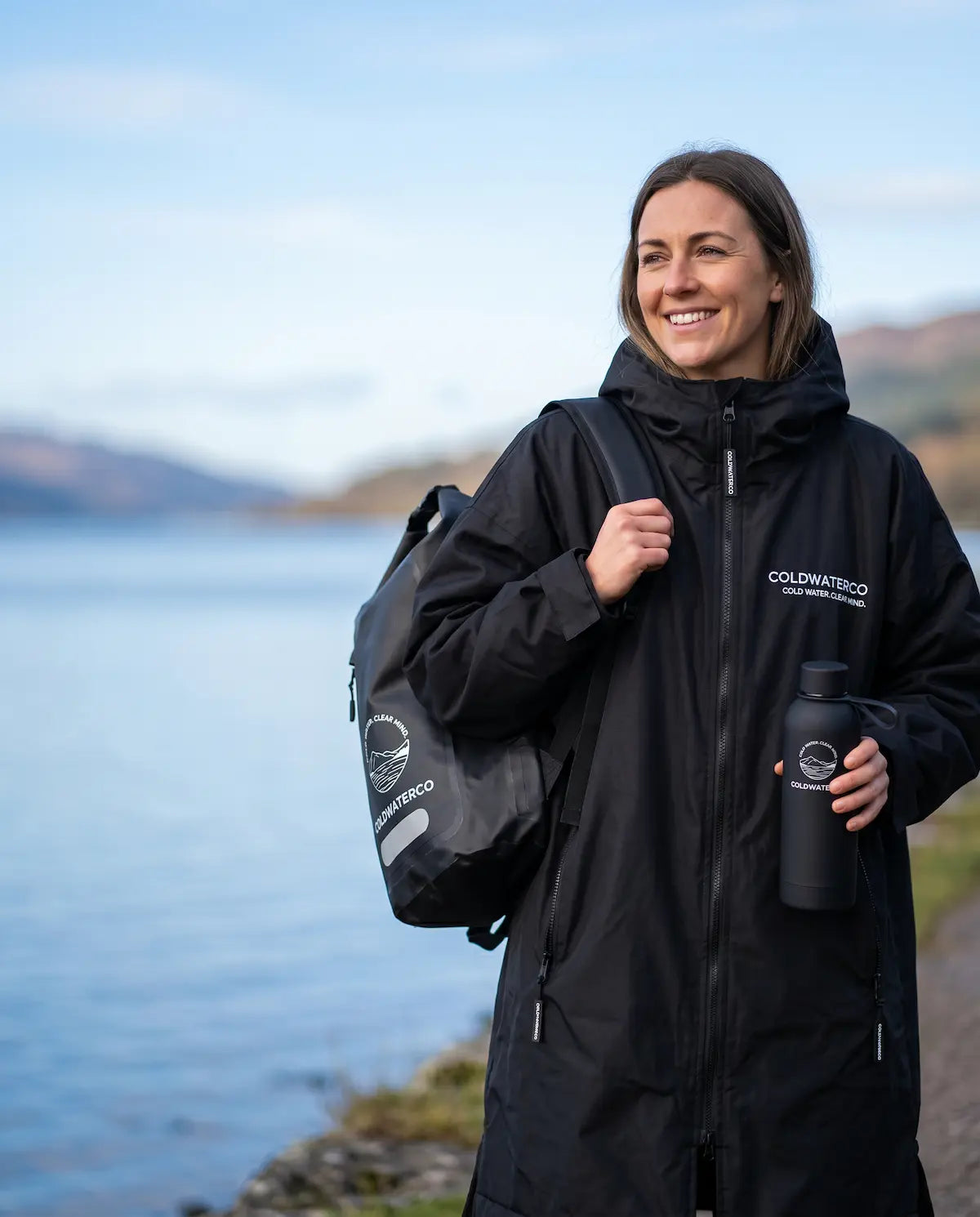 Person wearing a black jacket with 'COLDWATERCO' branding, holding a backpack and bottle by a body of water.