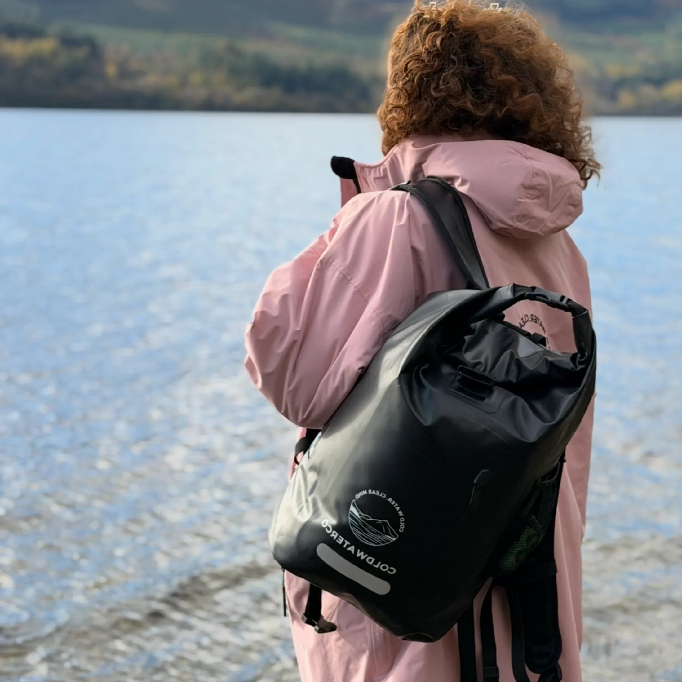 Person wearing a pink coat and carrying a black backpack by a lake with mountains in the background