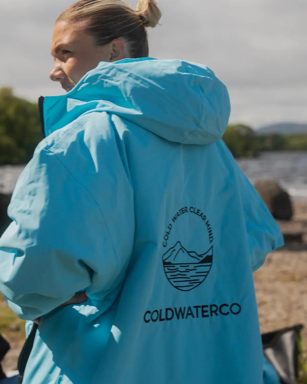 A woman wearing a bright light blue hooded jacket outdoors, photographed from the back. The jacket has a black circular mountain-and-water logo with the text 'COLDWATER CLEANING' and 'COLDWATERCO' printed on the back.