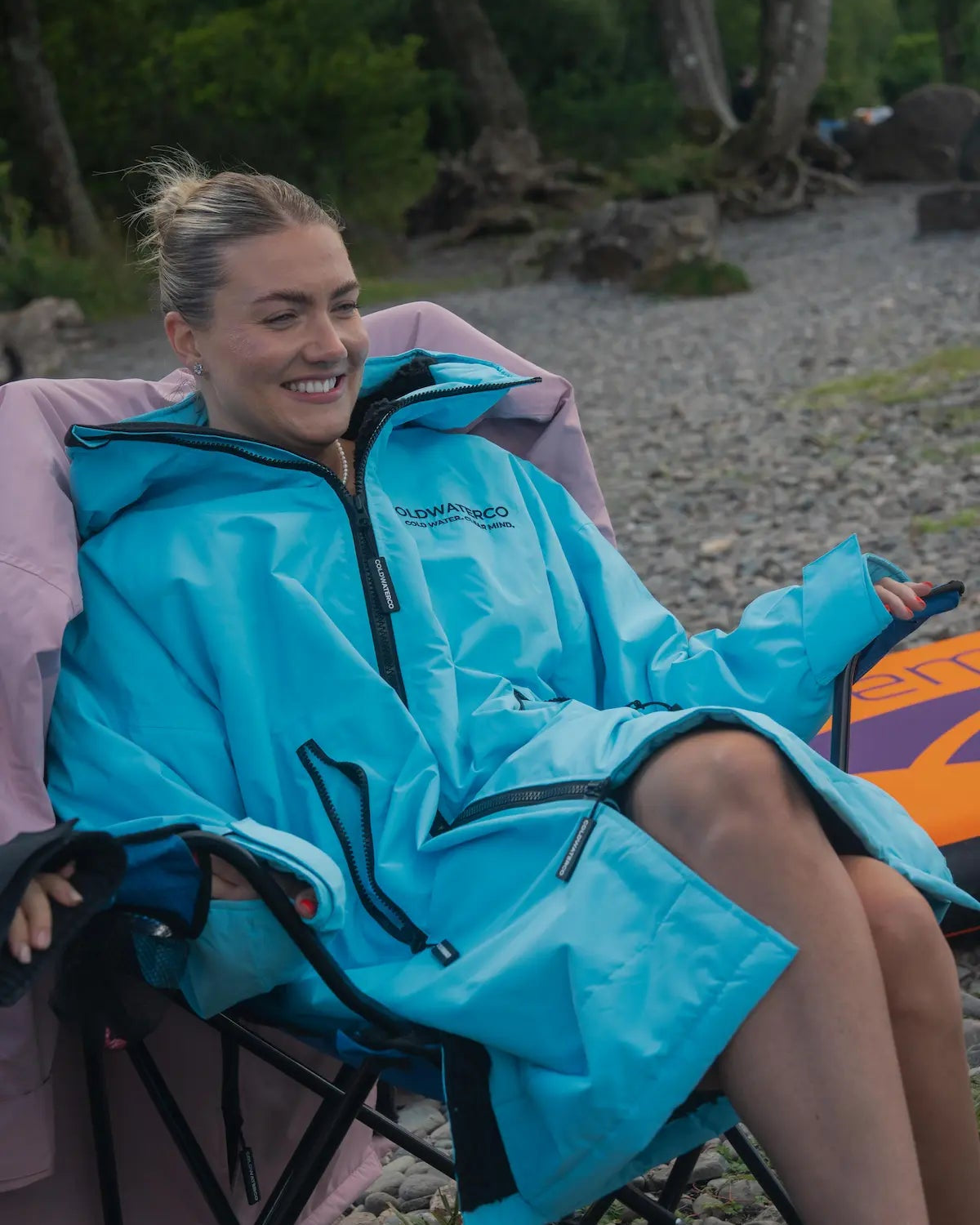 A woman sitting in a camp chair outdoors wearing a bright turquoise waterproof COLDWATERCO jacket/robe with black trim. The setting appears to be a rocky lakeside or forested outdoor area.