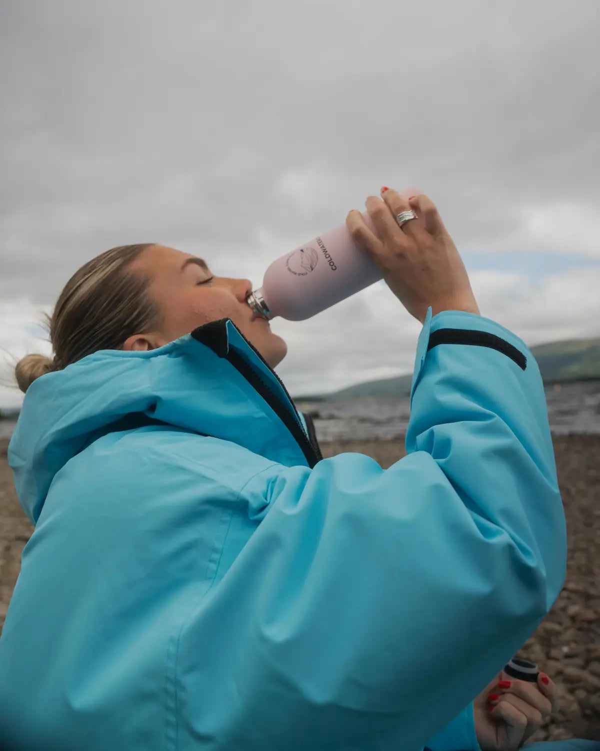 A woman in a bright turquoise COLDWATERCO jacket drinking from a light pink reusable COLDWATERCO bottle on a rocky beach under an overcast sky.