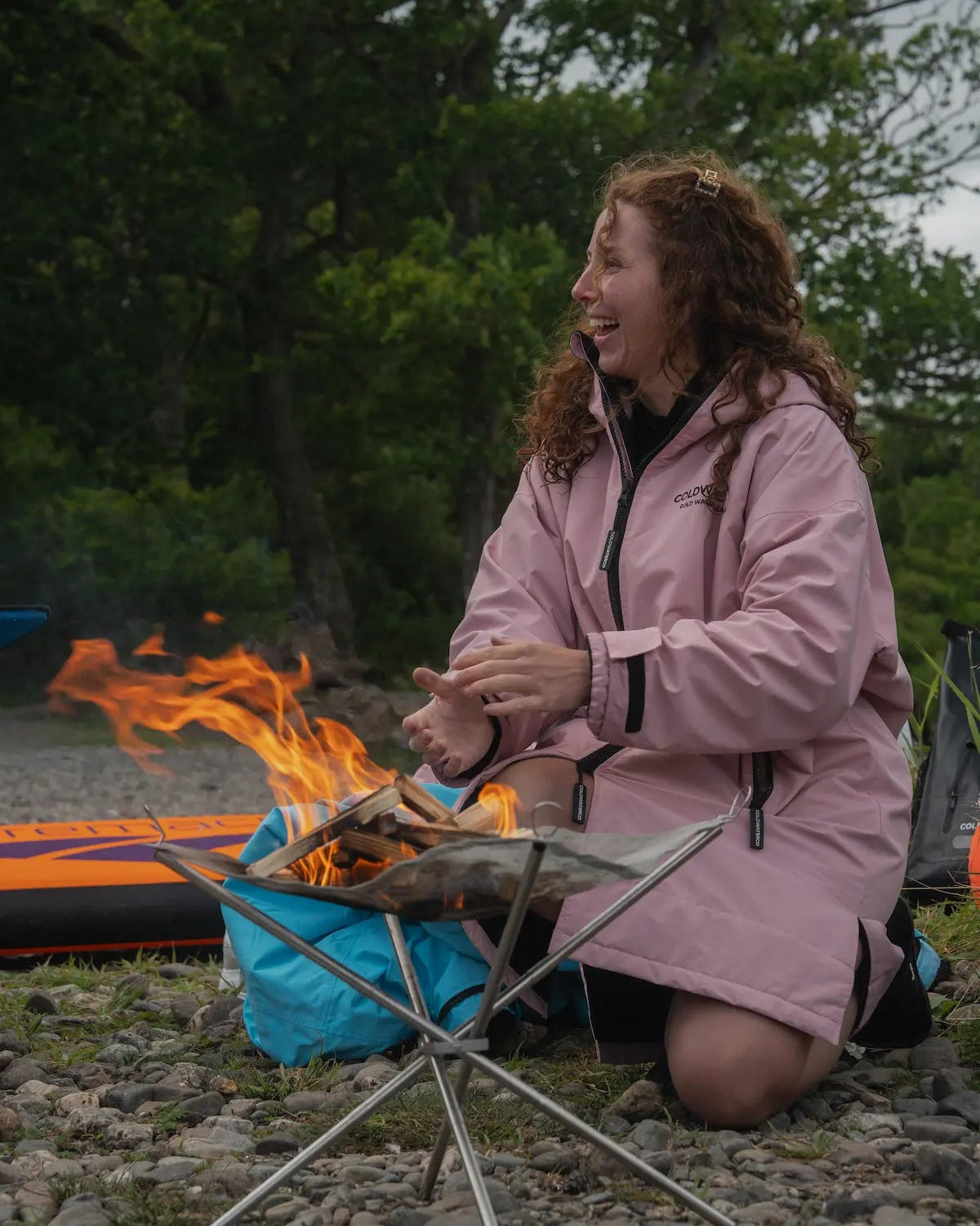 A woman kneels beside a small campfire on a rocky ground, wearing a light pink COLDWATERCO waterproof jacket. A blue bag, folding camp table, and part of an orange sleeping bag are visible in an outdoor wooded setting.