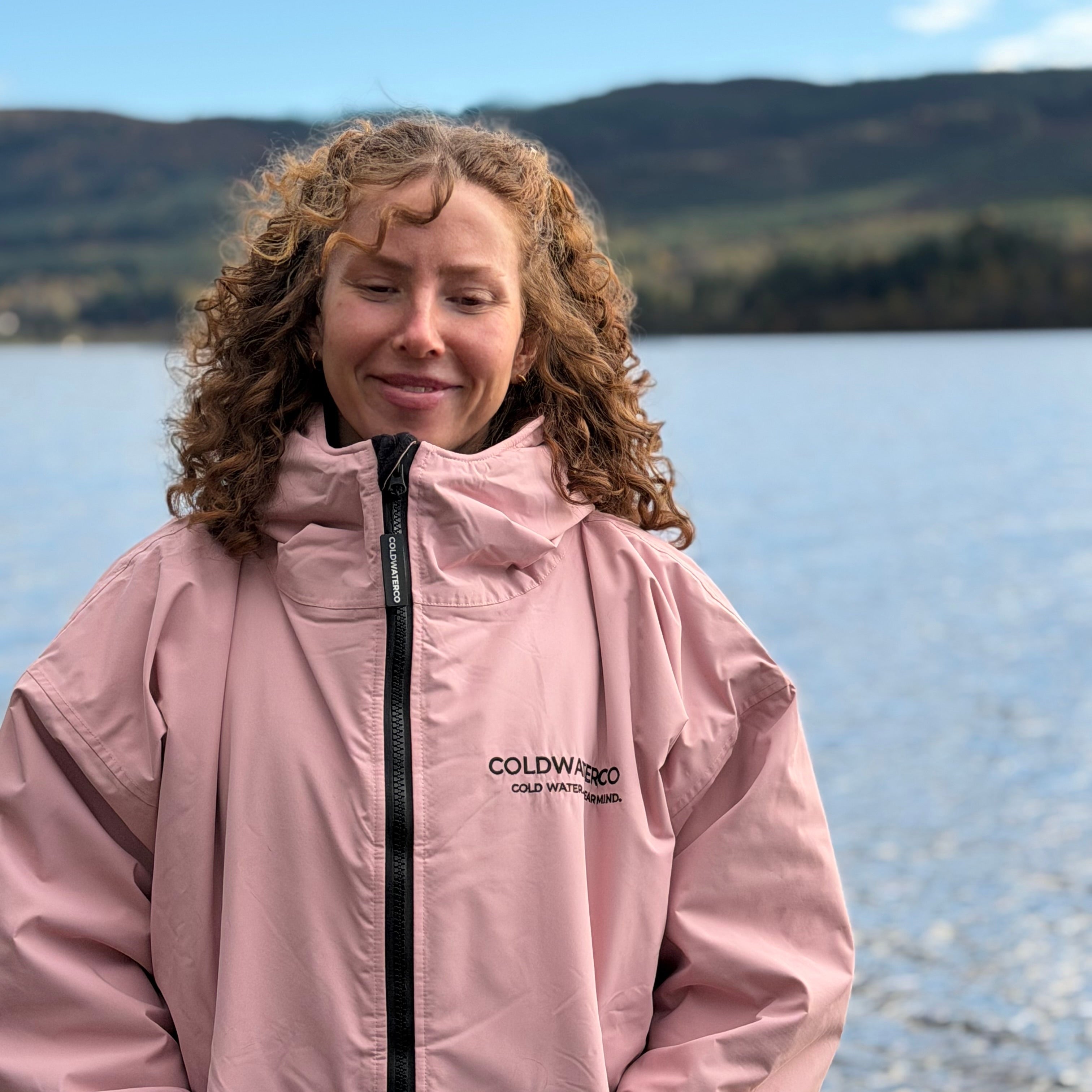 Person wearing a pink jacket with 'COLDWATER' branding by a loch with mountains in the background