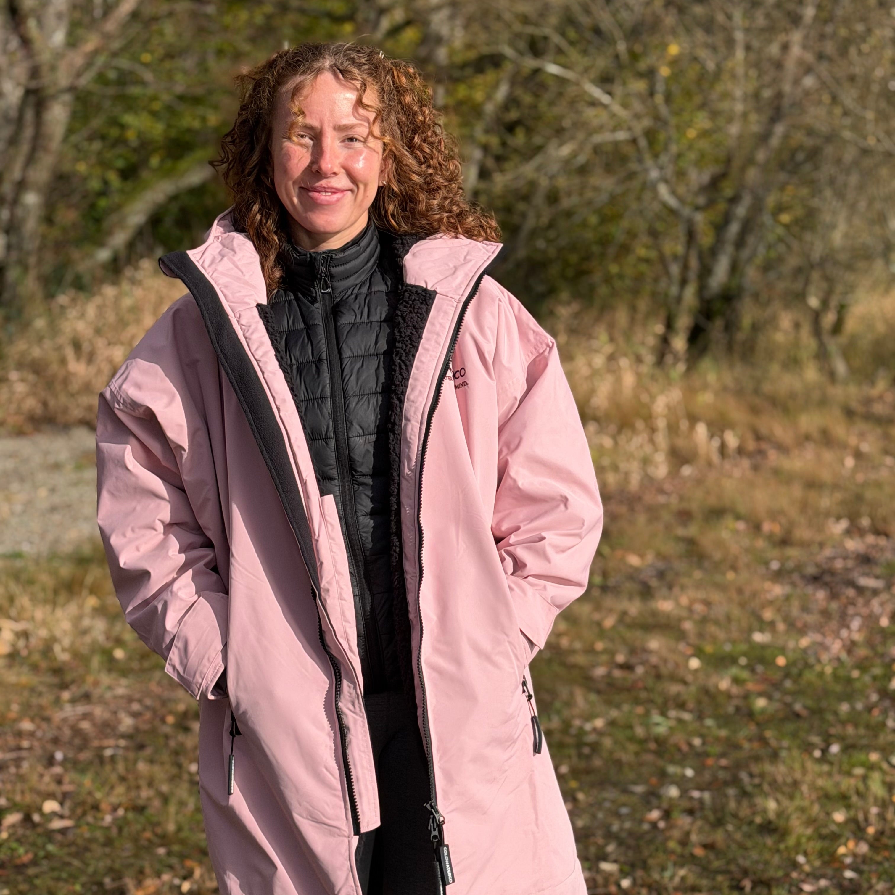 Person wearing a pink coat standing in a natural setting with trees and grass.