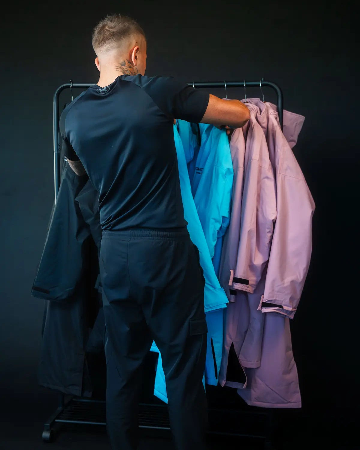 A man viewed from behind stands at a clothing rack, holding a dark ColdWaterCo Changing Robe jacket while reaching toward bright blue and lavender jackets hanging on the rack against a dark studio background.