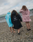 Three people walking barefoot on a rocky lakeshore, each wearing a hooded raincoat in different colors: light blue, black, and dusty pink. The back of each coat features a circular logo with the brand name COLDWATERCO.