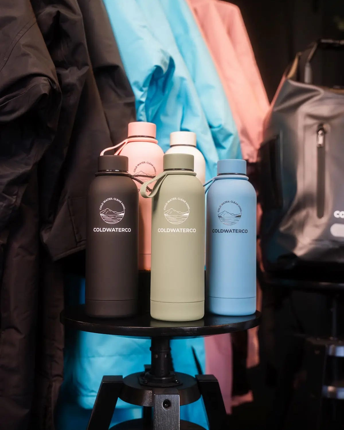 Five insulated water bottles on a black stool with colourful clothing in the background.