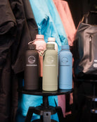 Five insulated water bottles on a black stool with colourful clothing in the background.