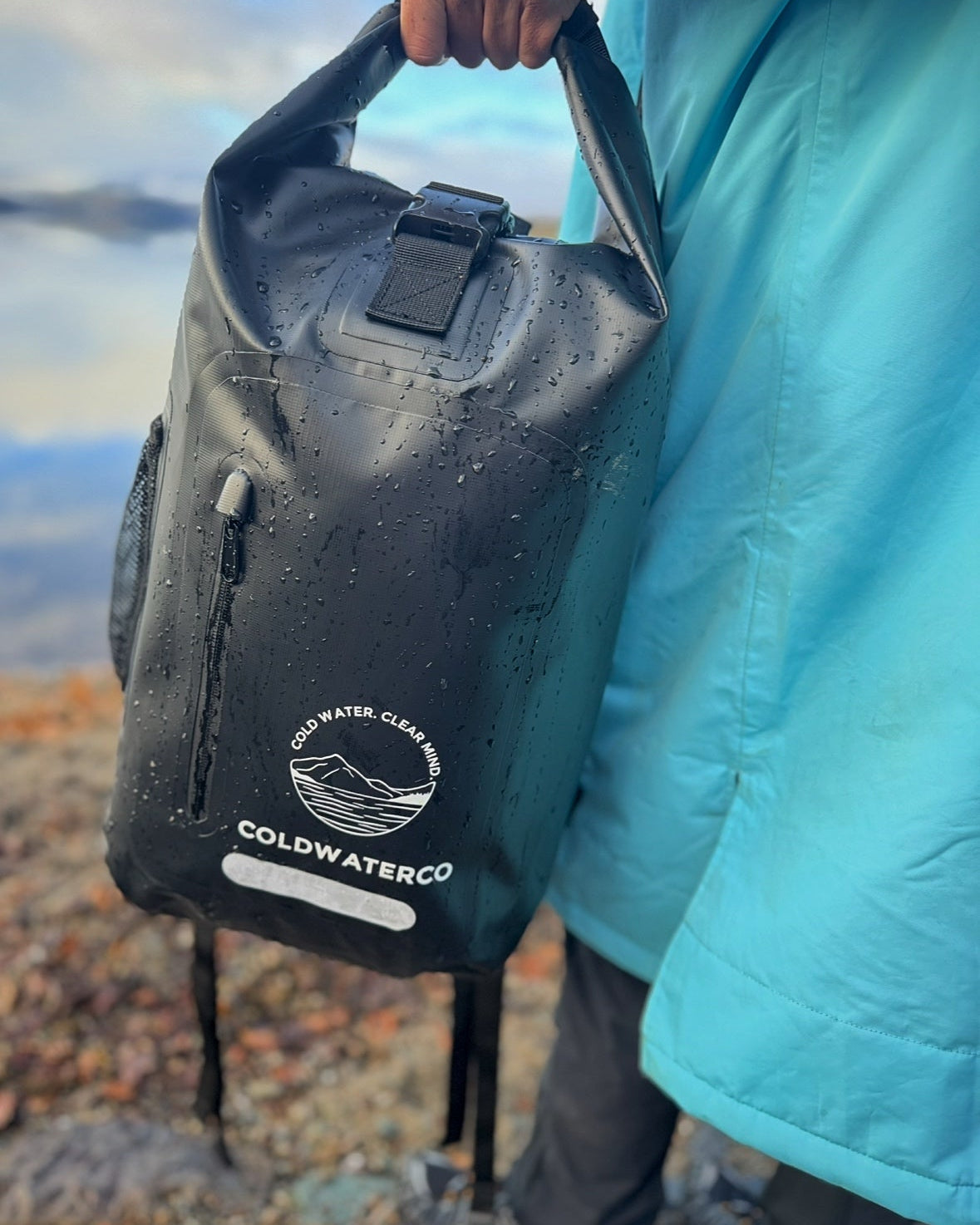 Black waterproof backpack shown in hand with water droplets on the bag. 