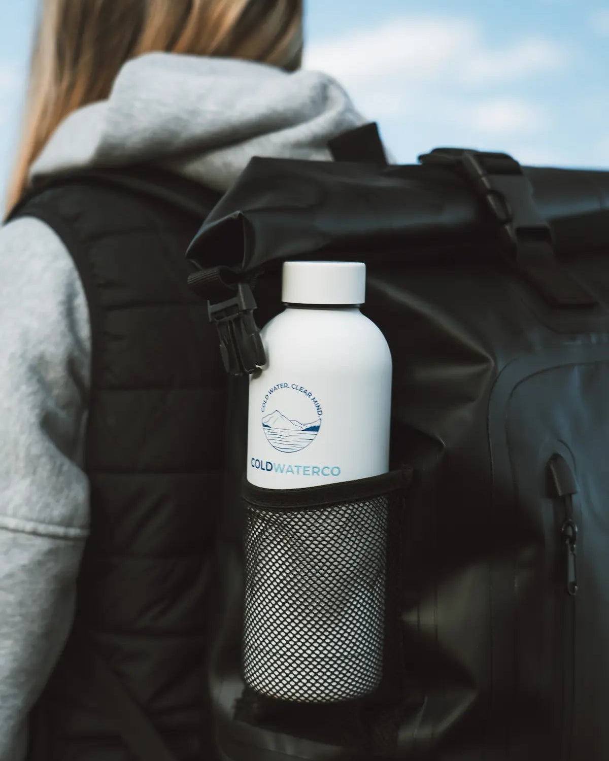 Person with a black backpack holding a white water bottle labeled 'Cold Water Co.' against a blue sky.