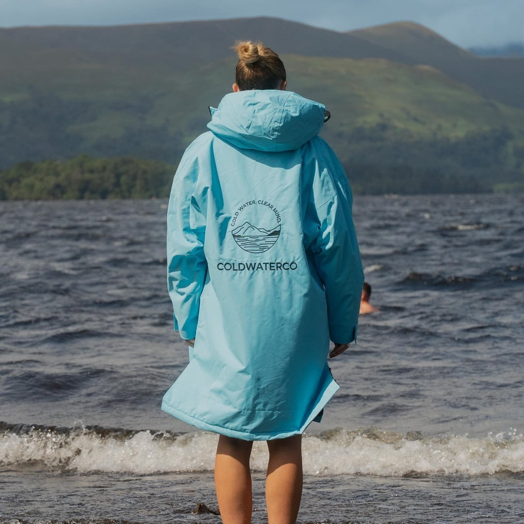 A person standing in shallow water at the beach, viewed from behind, wearing a light blue hooded raincoat with a circular logo and the word 'COLDWATERCO' printed on the back. Mountains and a cloudy sky are visible in the background.