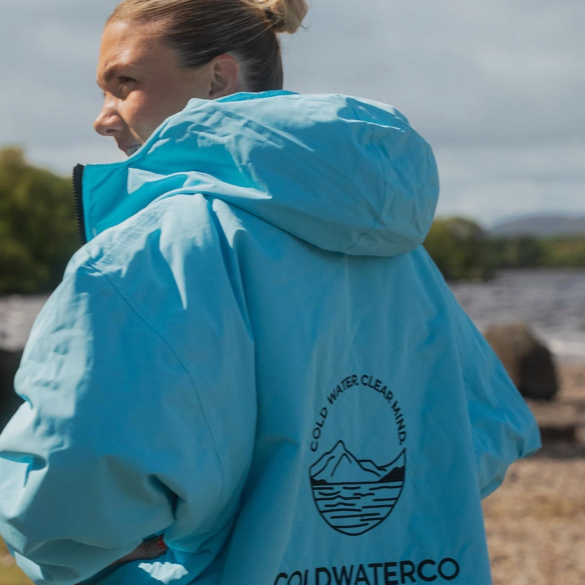 A woman wearing a bright light blue hooded jacket outdoors, shown from the back. The jacket has a black circular mountain logo with the text 'COLD WATER CLEAN MANNING' and 'COLDWATERCO' printed on the back.
