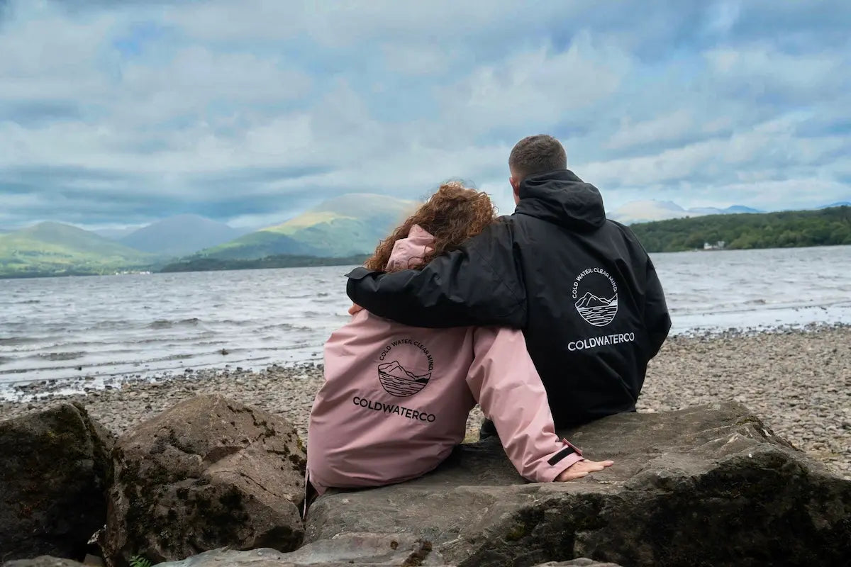 Two people sitting on rocks by a lake, seen from behind, wearing matching Coldwater Co hoodies and jackets. One person wears a black jacket and the other a pink jacket, both with the brand's circular mountain logo and 'COLDWATER CO' text on the back.