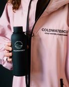 Person wearing a pink jacket with ColdwaterCo branding, holding a black water bottle.