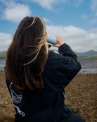 A woman with long brown hair with blonde streaks is seen from behind, wearing a black COLDWATERCO jacket with a white graphic on the back and holding a white COLDWATERCO bottle to her mouth. She is sitting outdoors on a rocky shore under a blue sky with clouds.