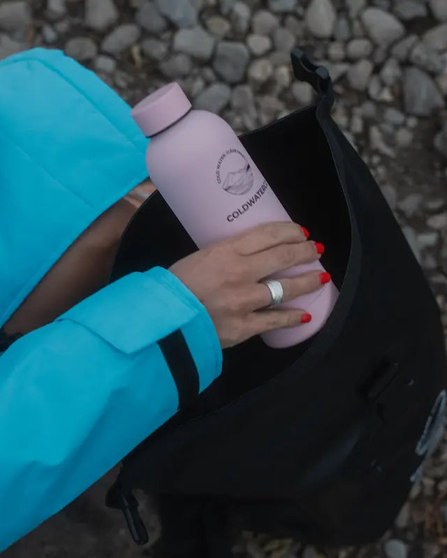A person in a bright turquoise COLDWATERCO jacket places a light pink insulated bottle into a black backpack, set against a background of gray pebbles.