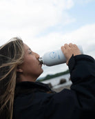 A woman drinking from a white reusable COLDWATERCO water bottle with a blue logo, set against an outdoor background with water and sky.