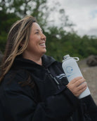 A woman wearing a black zip-up COLDWATERCO jacket/robe and holding a white reusable COLDWATERCO water bottle outdoors.