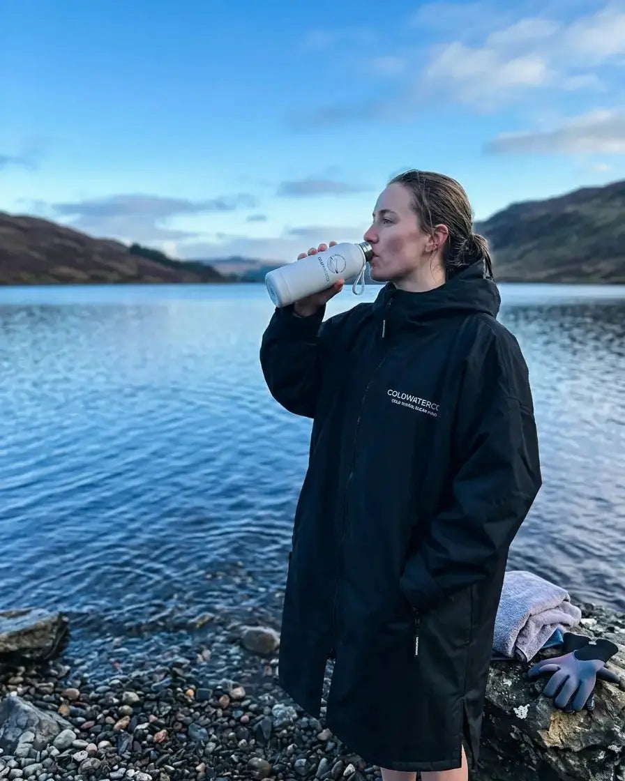 Person drinking from an insulated 'ColdWaterCo' water bottle by a loch with mountains in the background