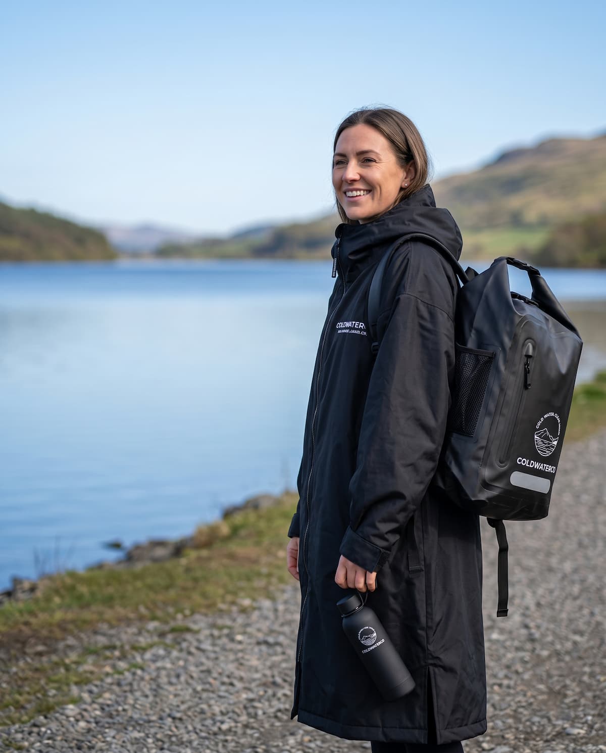 Person wearing a black 'ColdWaterCo changing robe and carrying a black backpack by a lake
