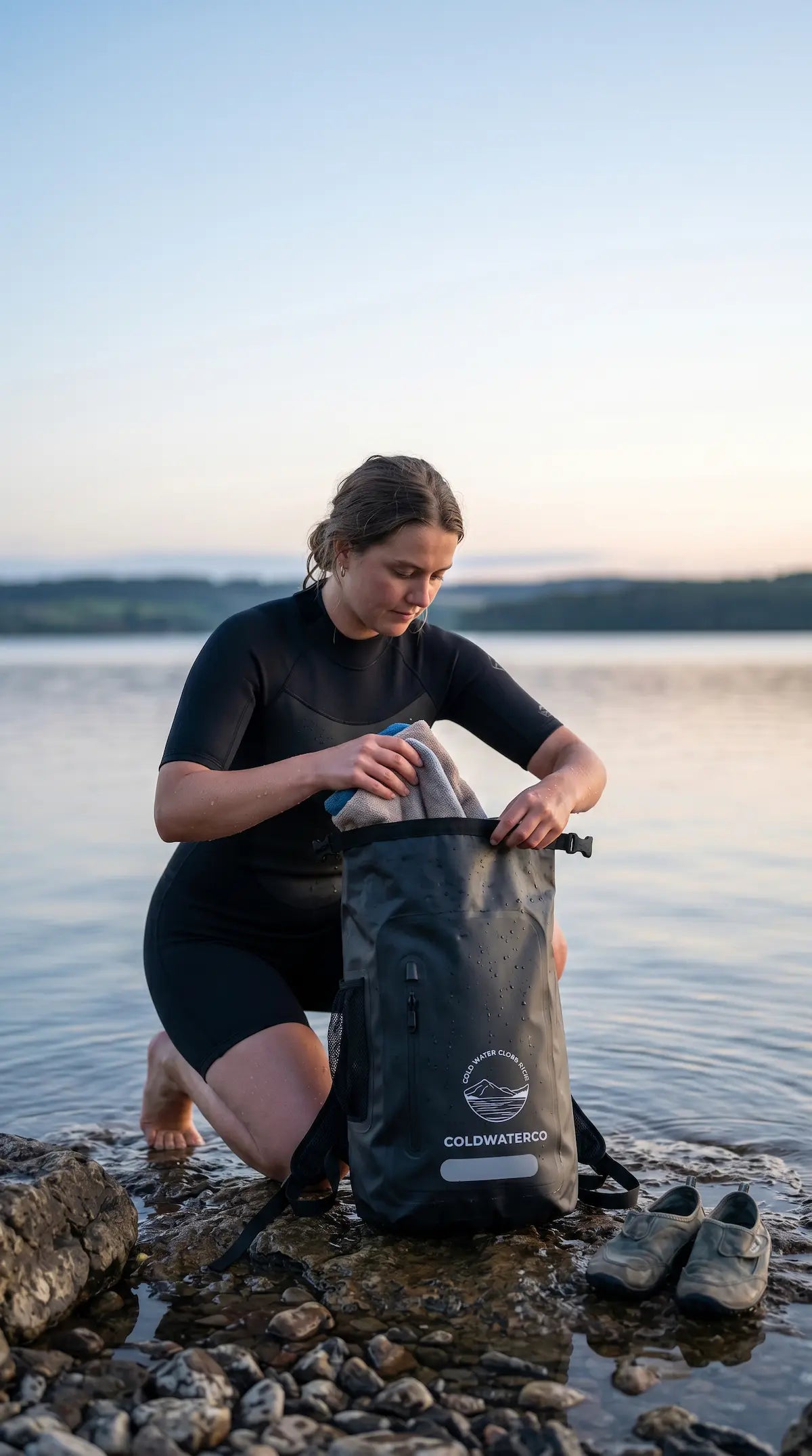 Person in a wetsuit opening a waterproof bag by a lake at sunset