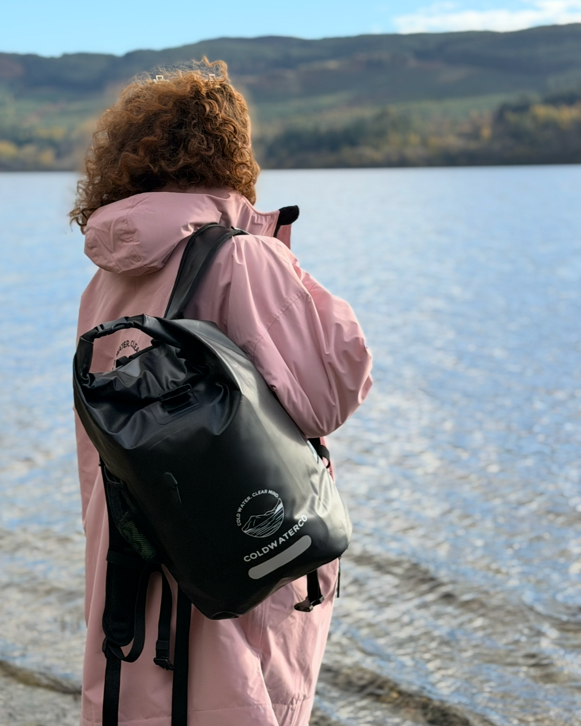 Person wearing a pink coat and carrying a black backpack by a lake with mountains in the background