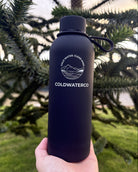 Black water bottle with 'COLDWATERCO' logo held in front of green foliage