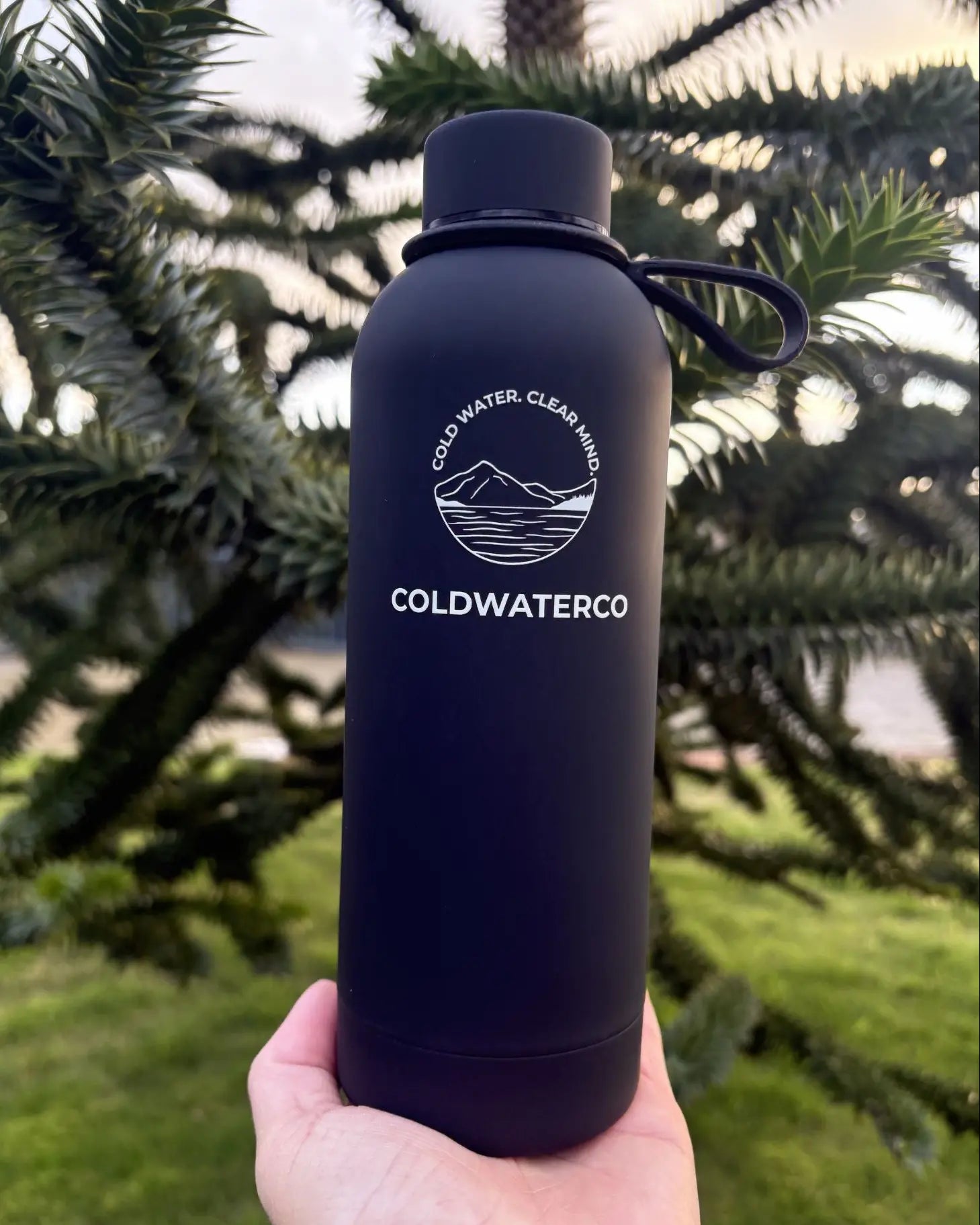 Black water bottle with 'Coldwaterco' logo held in front of a tree