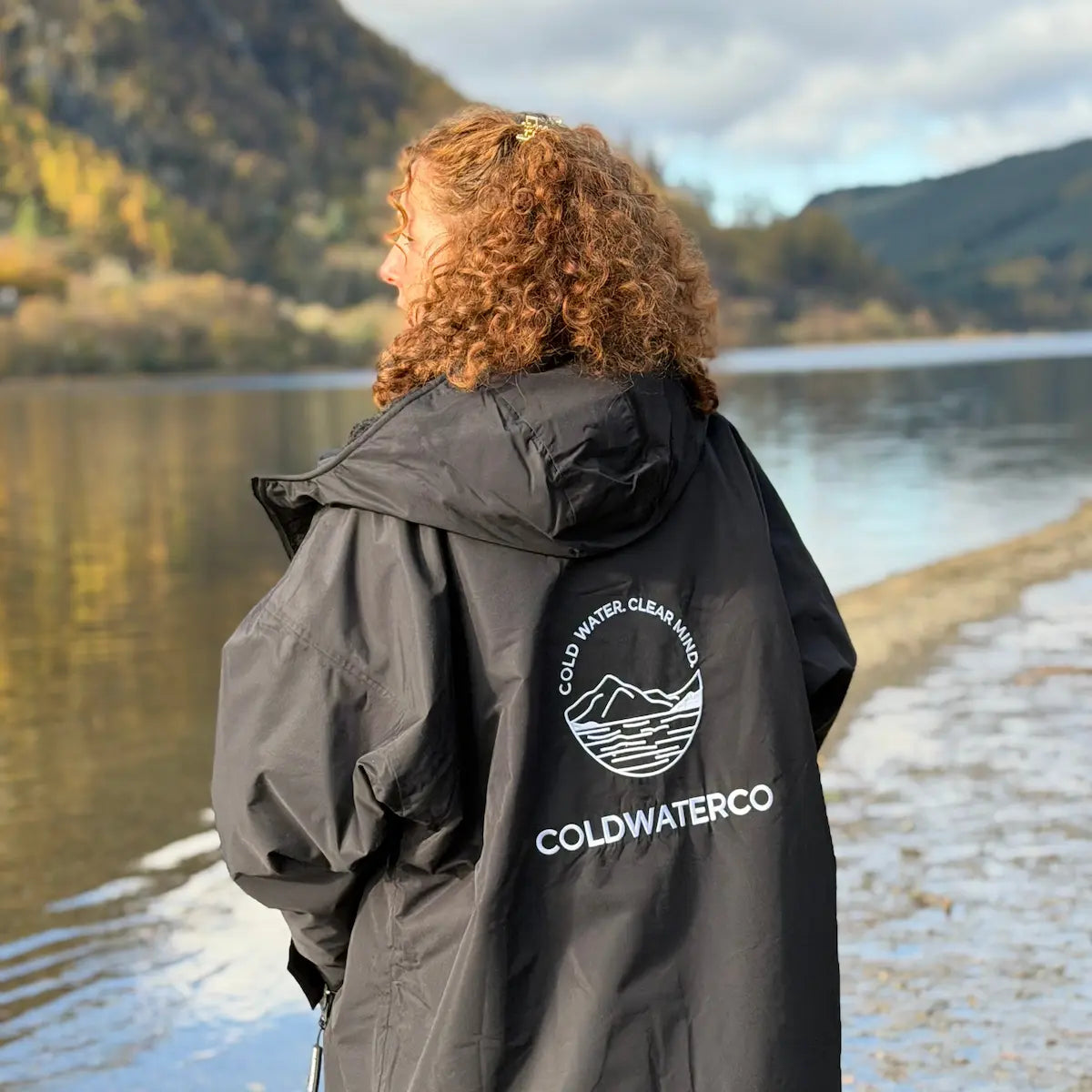 Person wearing a black jacket with 'Coldwater Co' logo by a loch