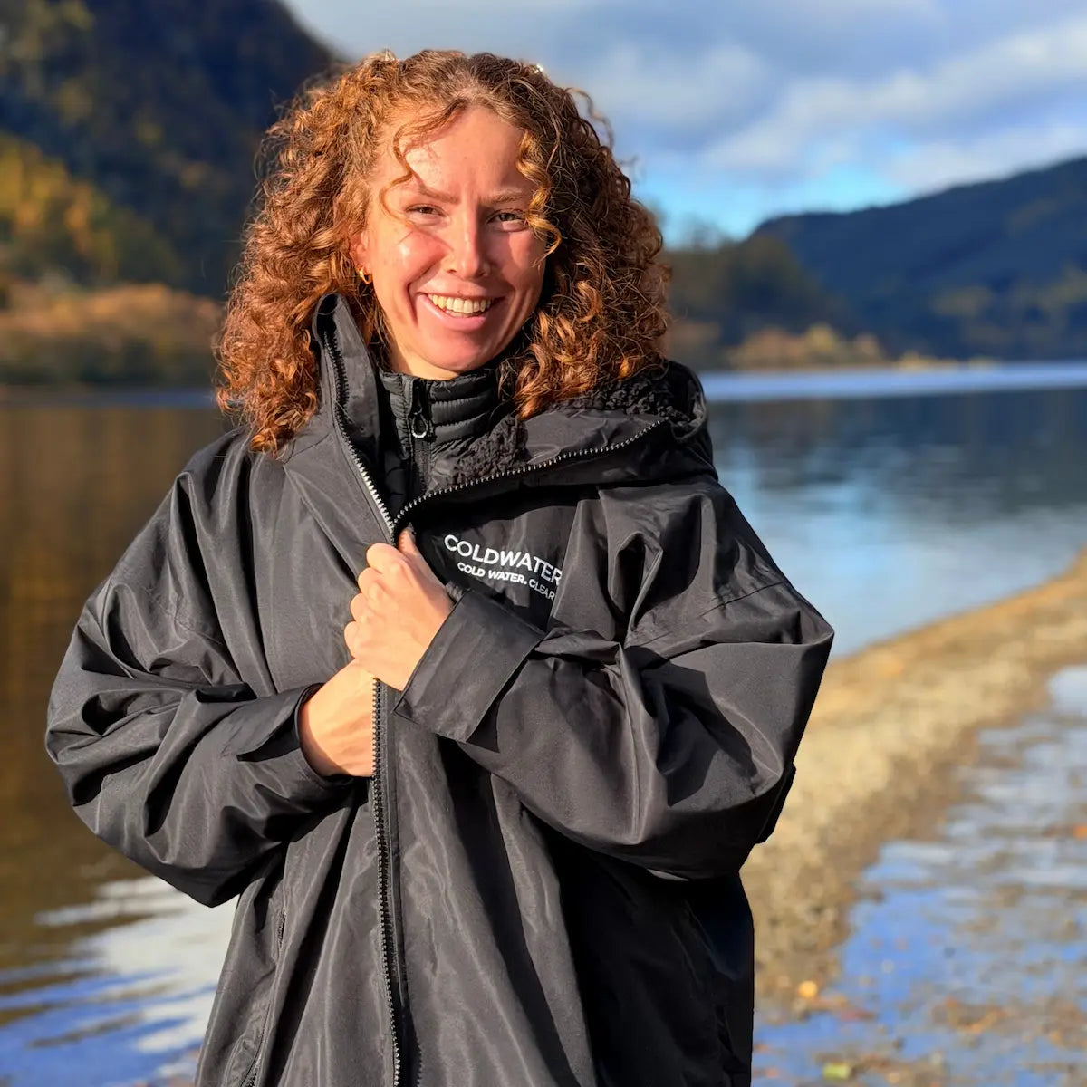 Person wearing a black jacket with 'Coldwater' branding by a lake with mountains in the background