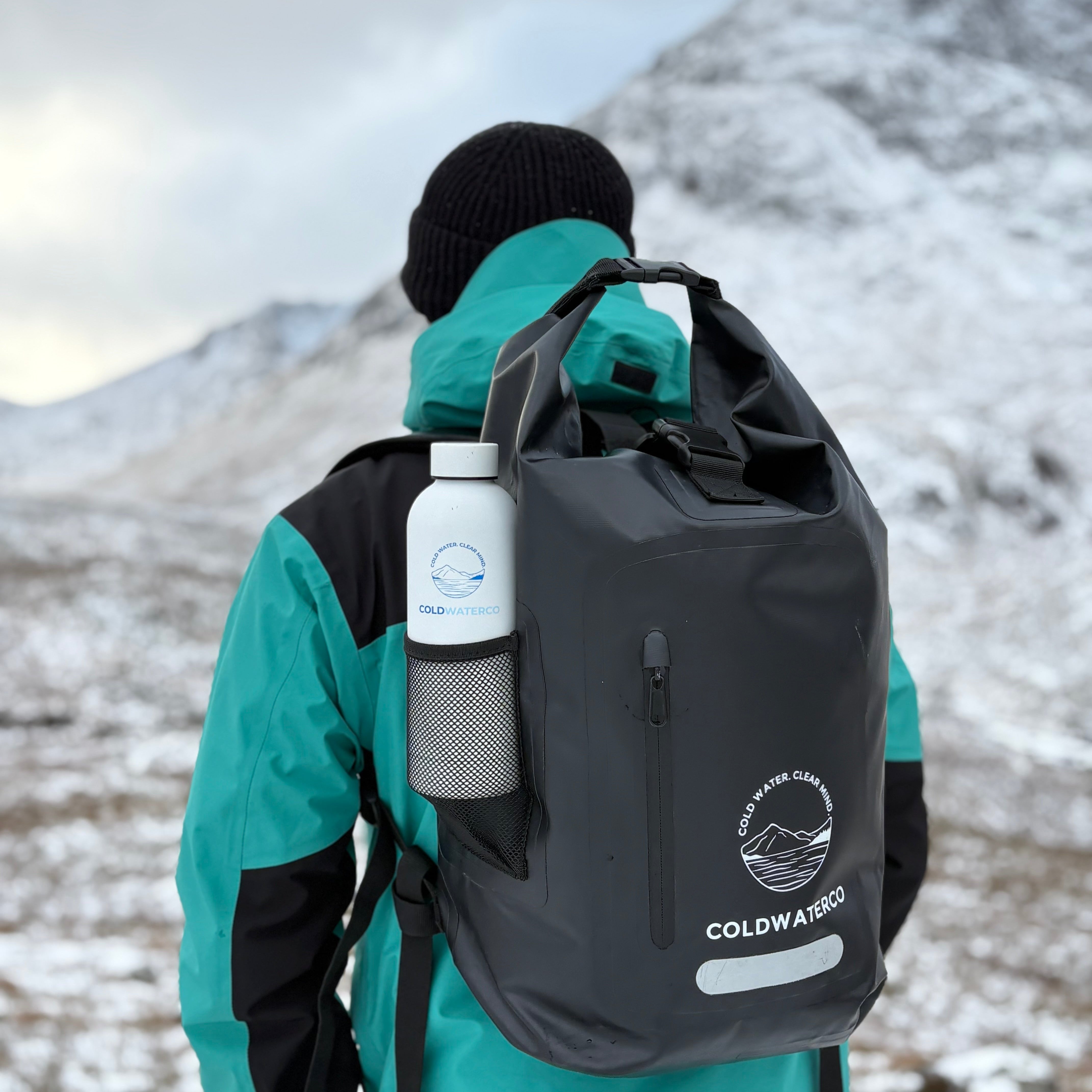 Person wearing a teal jacket and black backpack with 'Coldwater' branding in a snowy mountainous area.