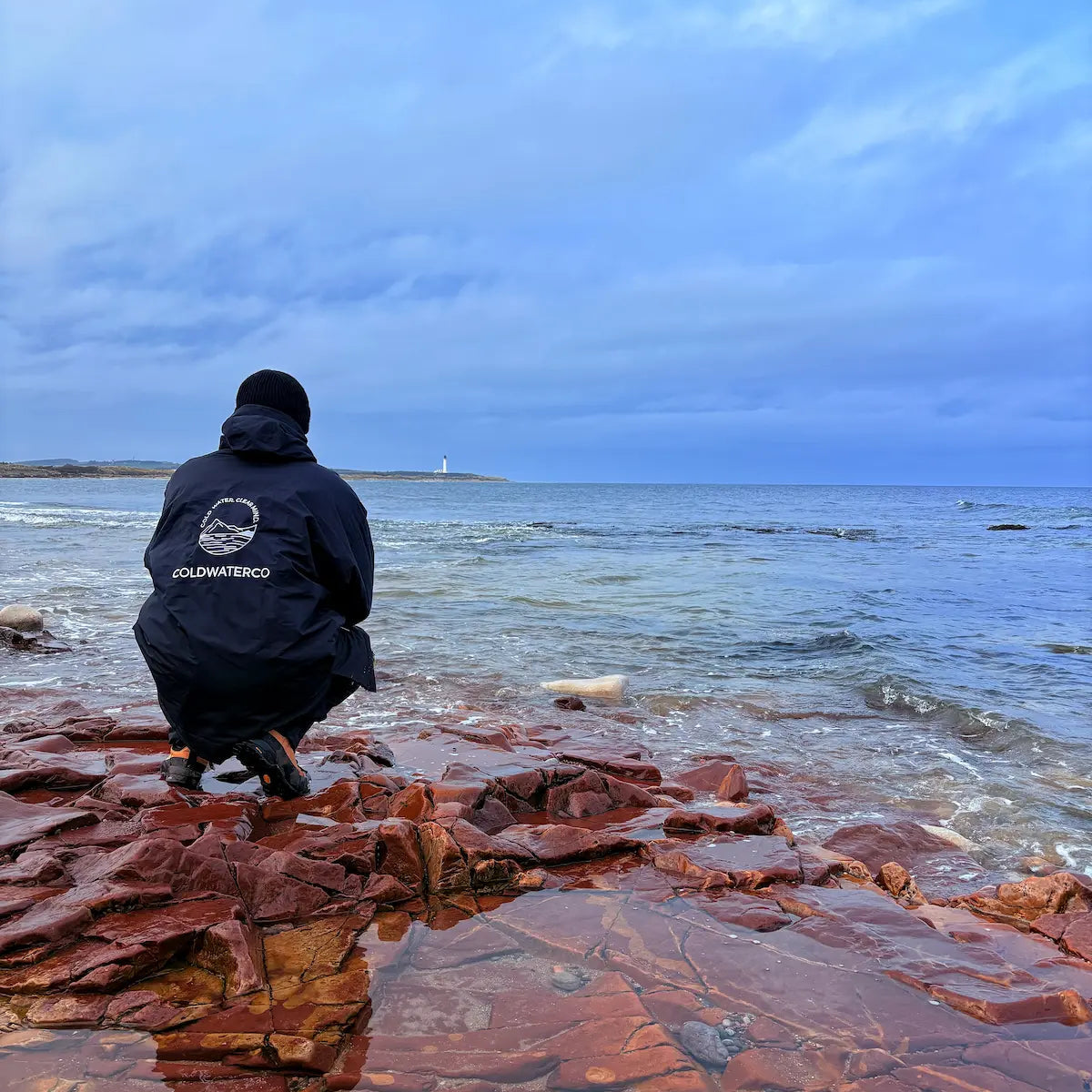 PErson crouched down at water edge with ColdWaterCo robe on