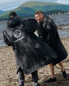 Two people wearing black waterproof poncho-style raincoats on a pebble beach with water and green hills in the background. One person faces away, showing a white circular logo and the word COLDWATERCO on the back of the coat.