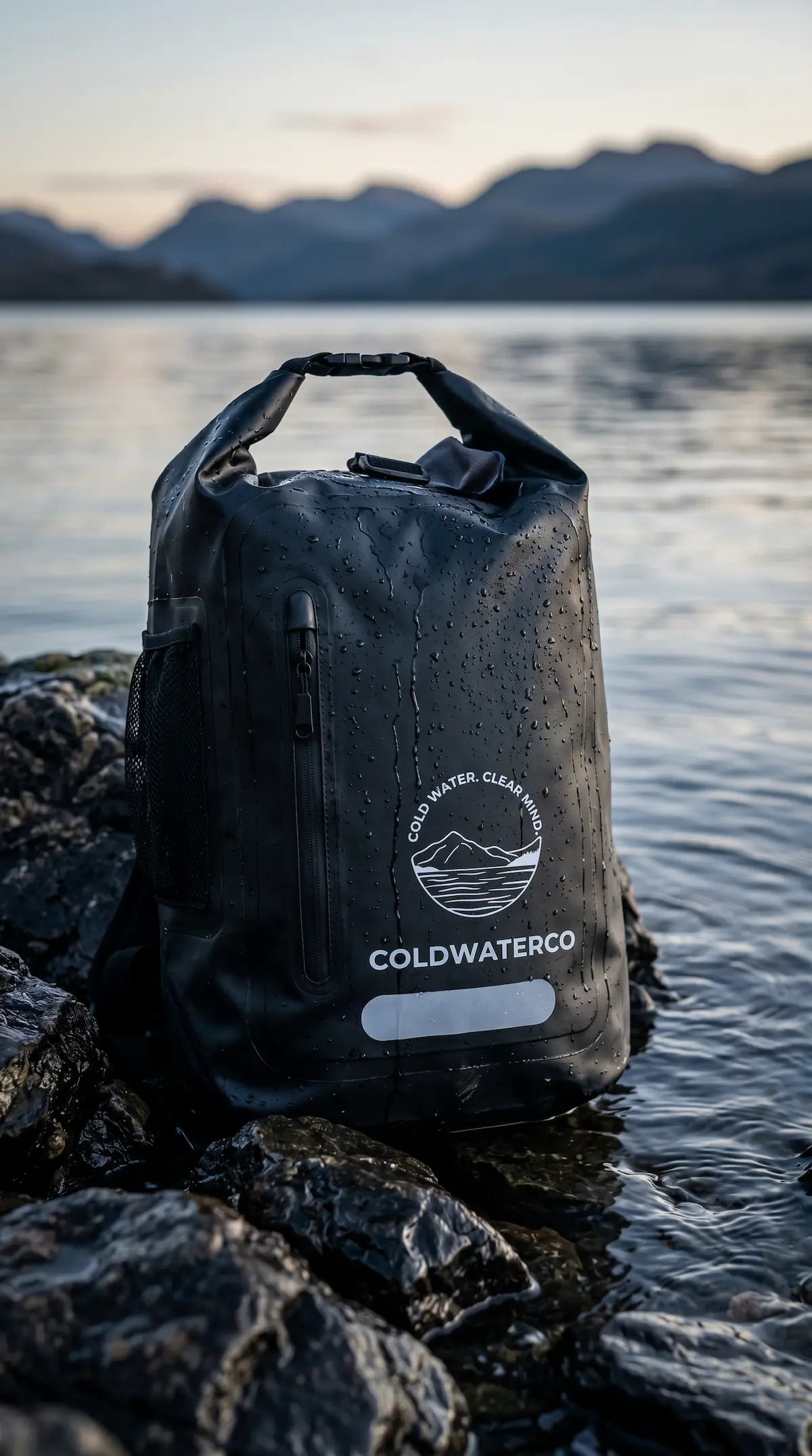 Black waterproof backpack with 'Coldwater Co' logo on rocks by a lake