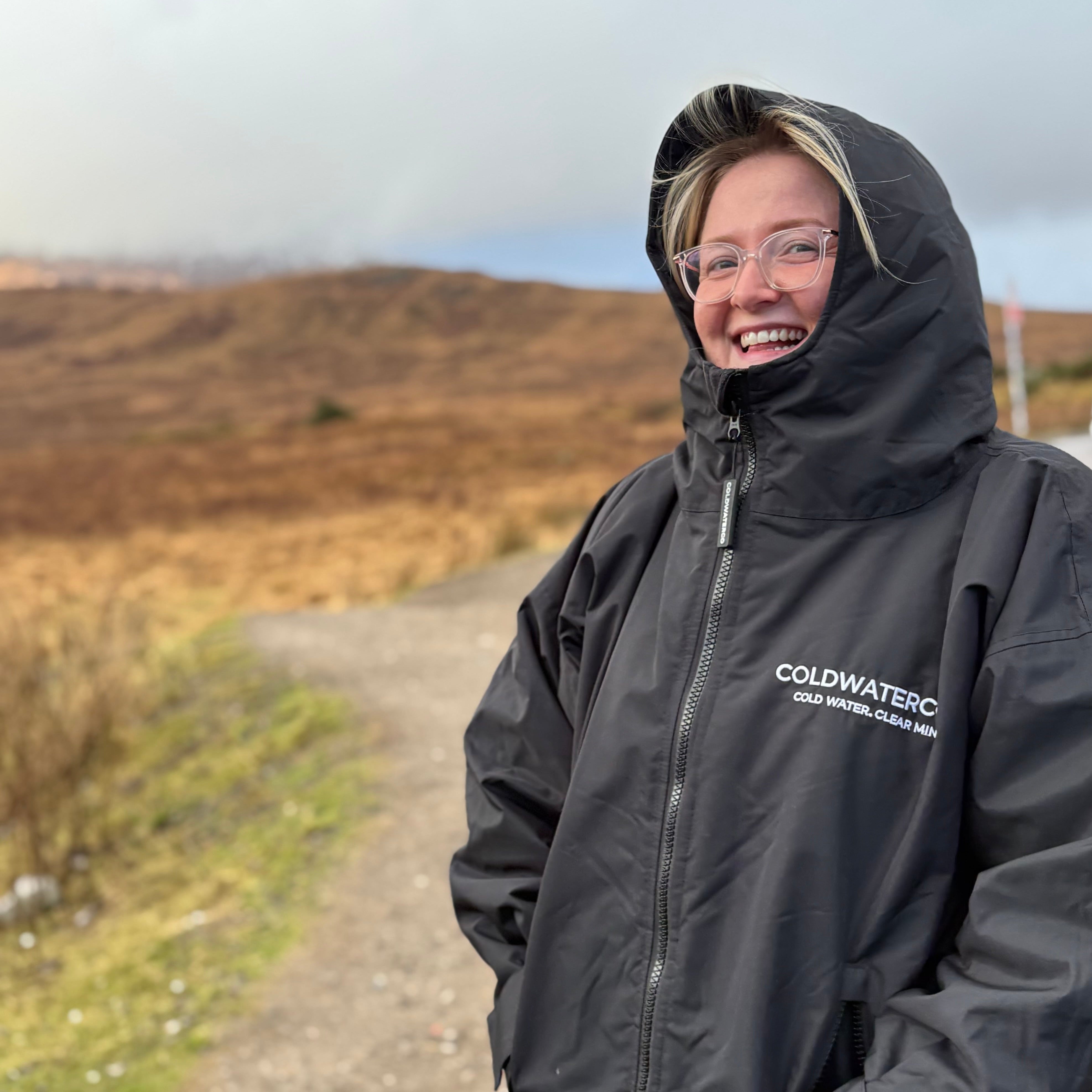 Person wearing a black ColdwaterCo jacket outdoors on a dirt road with mountains in the background.