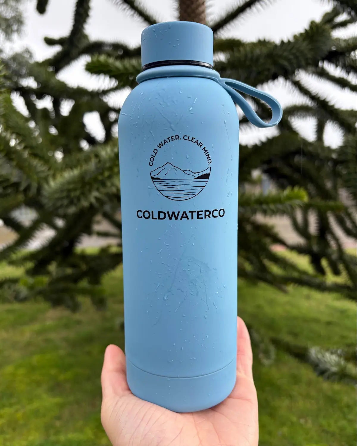 Blue water bottle with 'COLDWATERCO' branding held in front of a blurred natural background