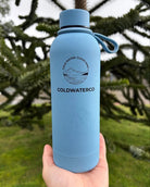 Blue water bottle with 'COLDWATERCO' branding held in front of a blurred natural background