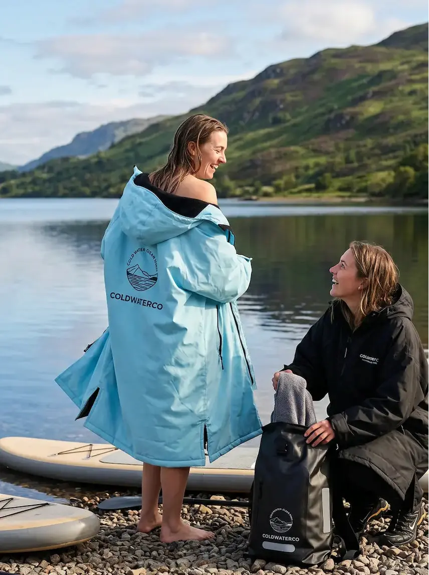 A woman wearing a light blue ColdWaterCO waterproof changing robe stands on a pebble shore by a lake, with a man kneeling beside her and paddleboards nearby. Mountains and water are visible in the background.