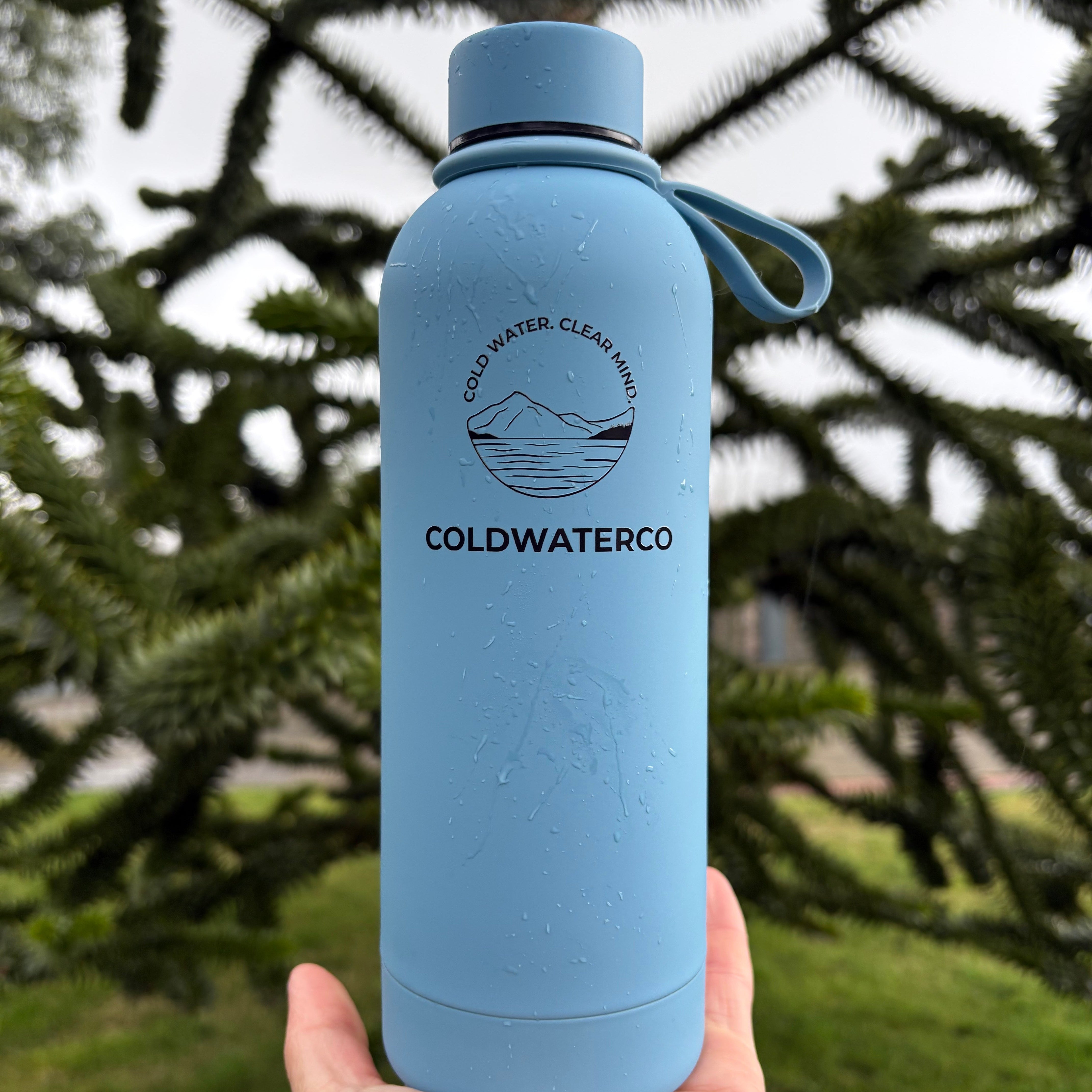 Hand holding a blue water bottle with 'COLDWATERCO' branding against a natural background.
