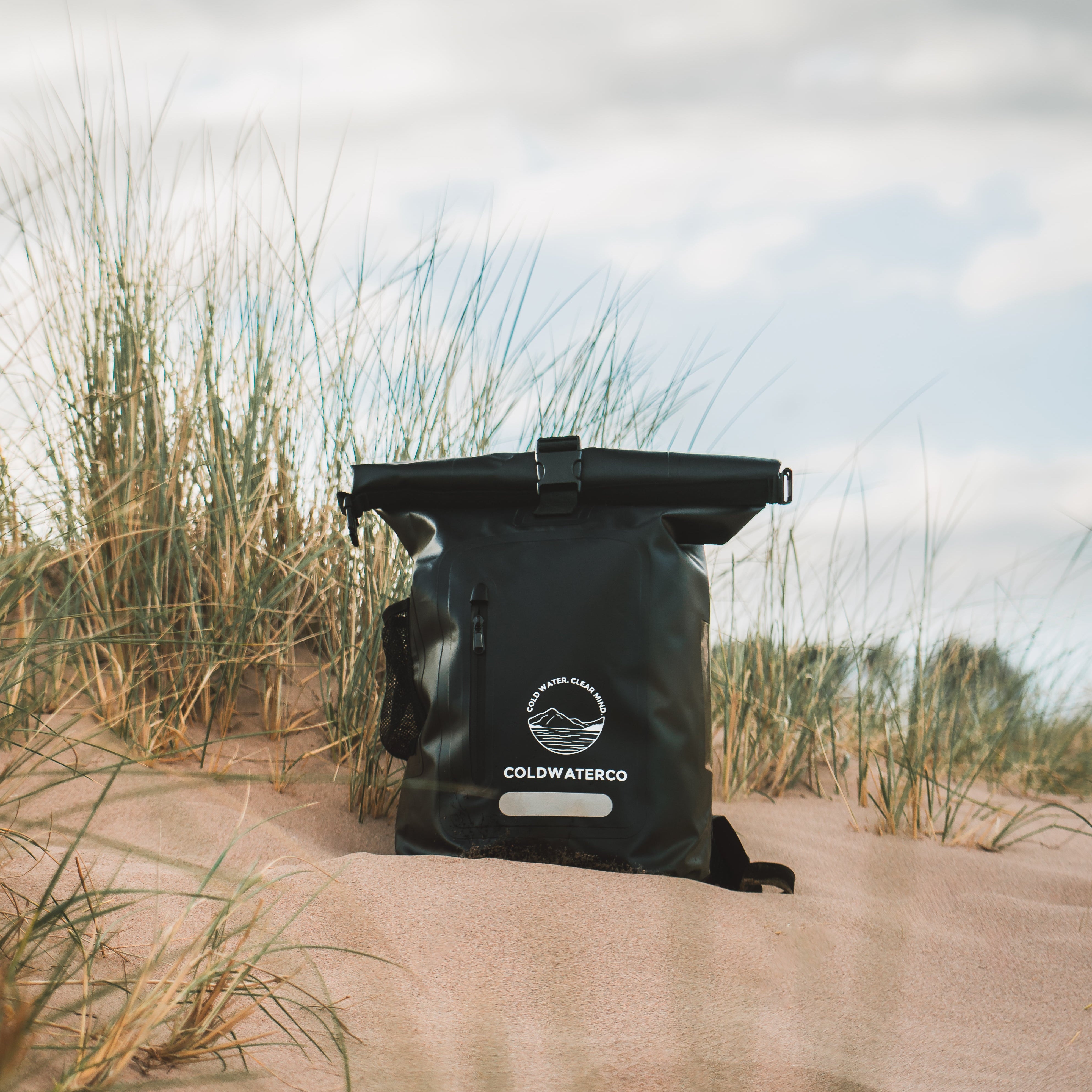 Black ColdWaterCo bag on a sandy beach with grasses and a cloudy sky.