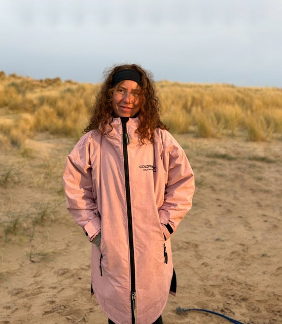 A woman with curly brown hair stands on a sandy beach wearing a light pink waterproof ColdWaterCo changing robe with black zippers and a black headband. The jacket has the brand name 'COLDWATERCO' printed on the left chest.