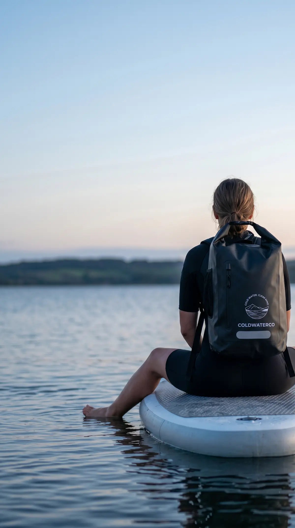 Person sitting on a paddleboard with a ColdWaterCo Outdoors backpack, facing a calm lake at sunset.