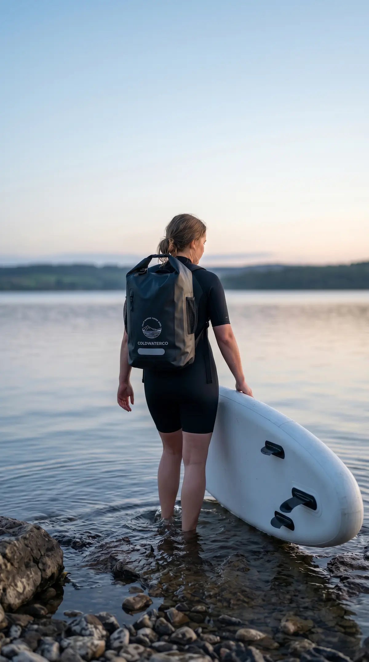 Person with a backpack and surfboard standing on a rocky shore by a body of water.