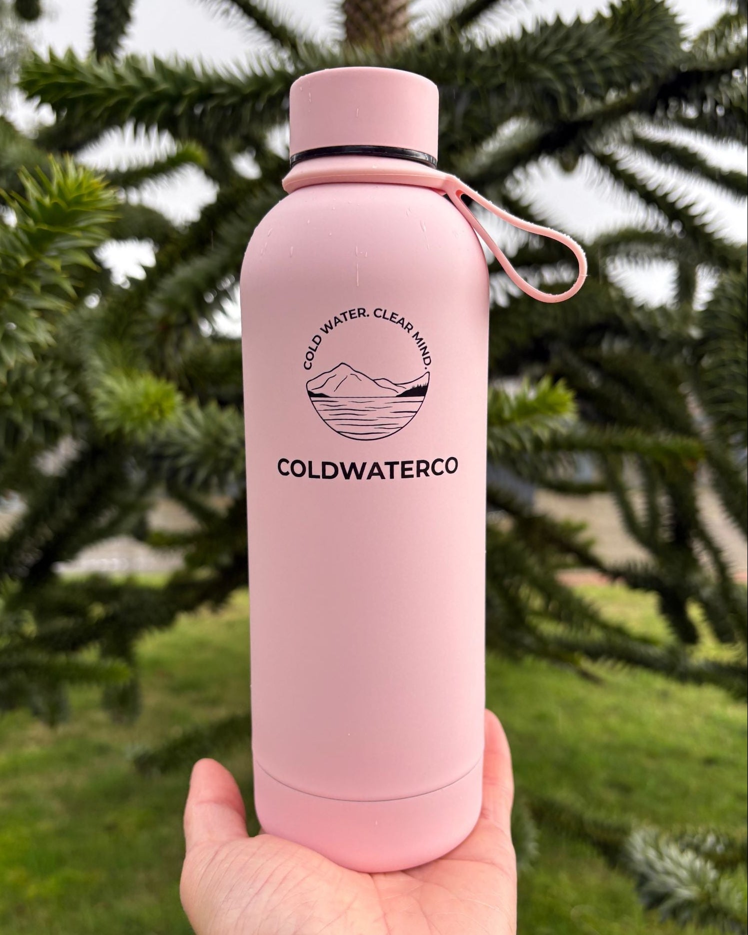 Pink water bottle with 'Coldwaterco' branding held in front of green foliage