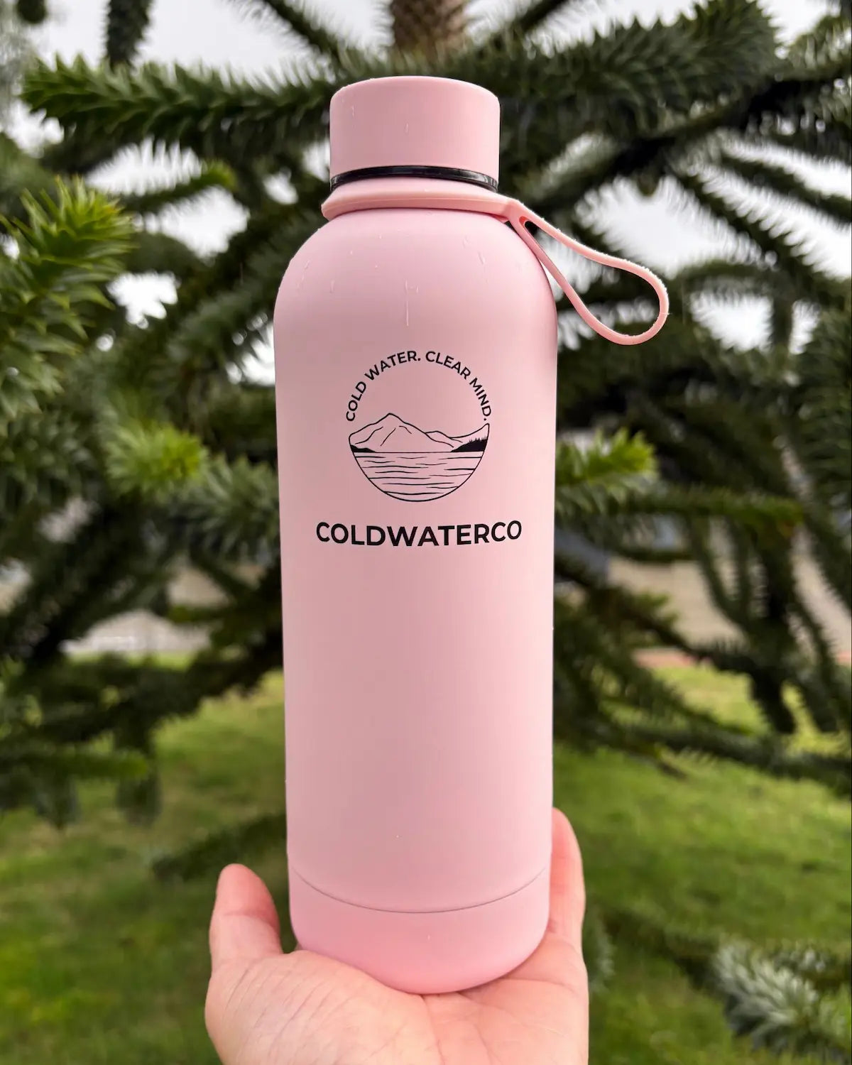 Pink water bottle with 'Coldwaterco' branding held in front of green foliage